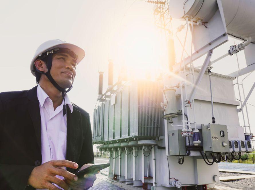 Businessman wearing a black suit, standing looking at a large power transformer with orange sunlight and white sky to be background, Concept about business people who want to invest in energy.